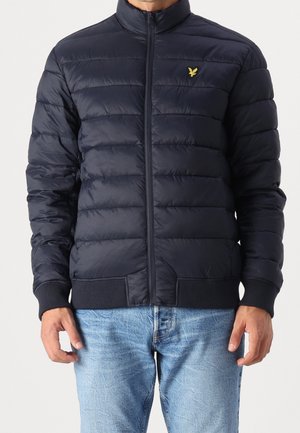 Homme portant une veste matelassée bleu marine zippée avec poignets et ourlet côtelés, associée à un jean bleu clair.