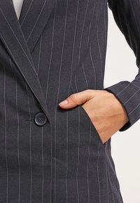 Blazer bleu marine à rayures fines avec fermeture à un bouton, revers crantés et poche latérale en tissu texturé.