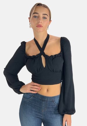 Schwarzes, kurzes Top mit langen Puffärmeln, rüschiertem Ausschnitt und einem frontalen Schlüsselloch-Ausschnitt. Kombiniert mit blauen Jeans.