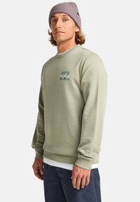 Lichtgroene sweatshirt met ribgebreide boorden en zoom, voorzien van een kleine golfafbeelding op de borst. Draagt over een wit shirt en gecombineerd met donkere broek.