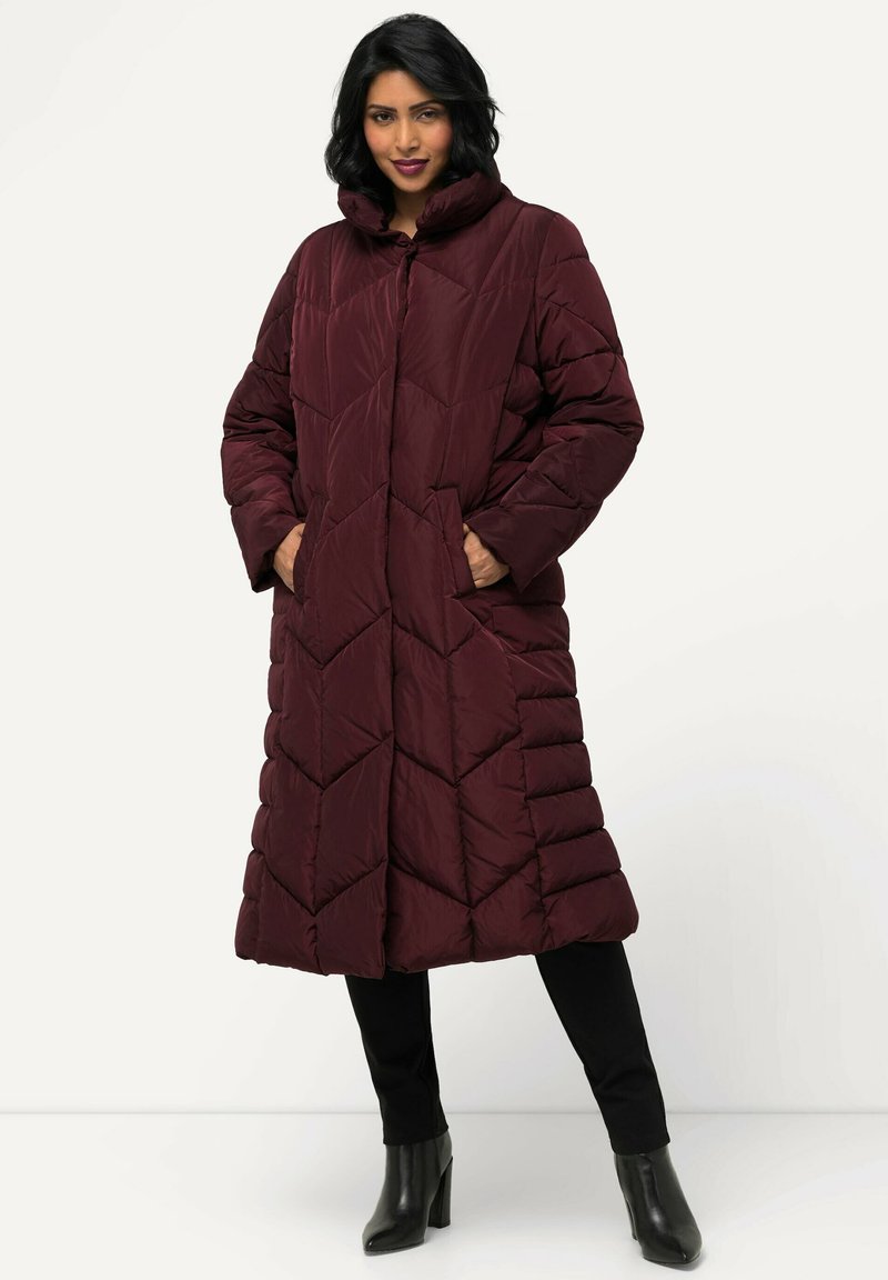 Cappotto imbottito bordeaux con collo alto, motivo a chevron trapuntato, due tasche laterali e design fino al ginocchio. Indossato sopra pantaloni neri e stivali.