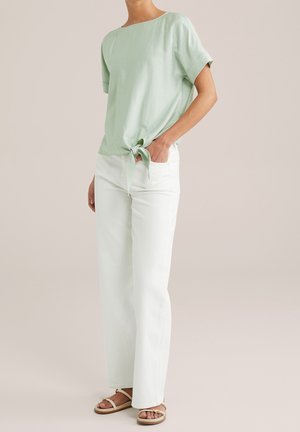 Persoon draagt een lichtgroene top met korte mouwen, gestrikt in de taille, witte broek met rechte pijpen en beige platte sandalen, staand tegen een effen achtergrond.