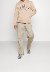 Sudadera rosa claro con logo negro de "JORDAN", pantalones de pierna ancha beige y zapatillas deportivas grises con acentos multicolores. Textura de tela suave.