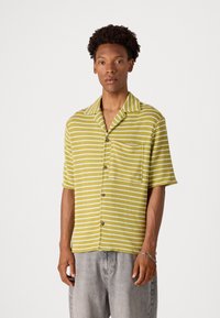 Chemise à manches courtes verte avec des rayures blanches, fabriquée en tissu texturé. Elle possède un col en V et une poche poitrine. Fermeture à boutons sur le devant.
