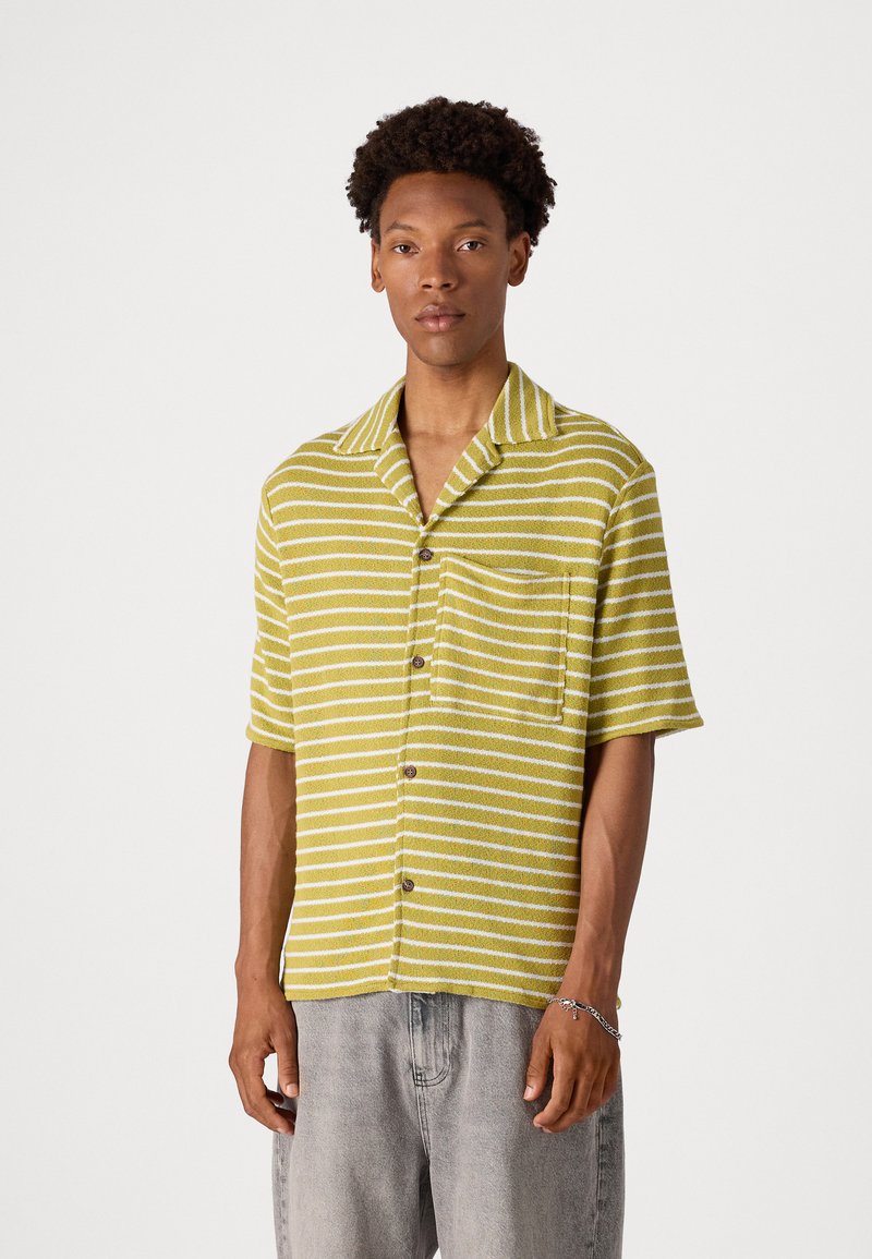 Chemise à manches courtes verte avec des rayures blanches, fabriquée en tissu texturé. Elle possède un col en V et une poche poitrine. Fermeture à boutons sur le devant.
