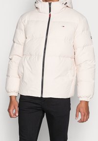 Pufferjack in lichtbeige, met een ritssluiting aan de voorkant, elastische manchetten en een capuchon met trekkoord. Logo detail op de borst, gladde textuur.