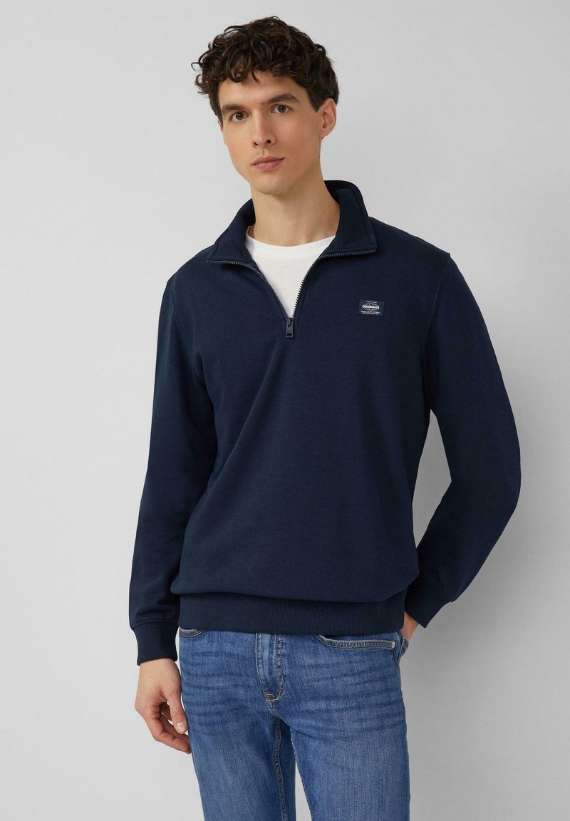 Vīrietis tērpies tumši zilā pusražotā džemperī uzvilktā virs baltas kreklas un zilām džinsām, stāv ar vienu roku kabatā vienkārša fona priekšā.