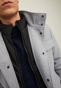 Grå ullblandad kappa med hög krage, med ett svart zippered innerlager och en texturerad marinblå tröja under. Knappar synliga på kappan.