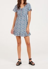 Robe fleurie bleue avec manches bouffantes, encolure en V et ourlet à volants. Le tissu présente de petites fleurs blanches et roses sur un fond uni.