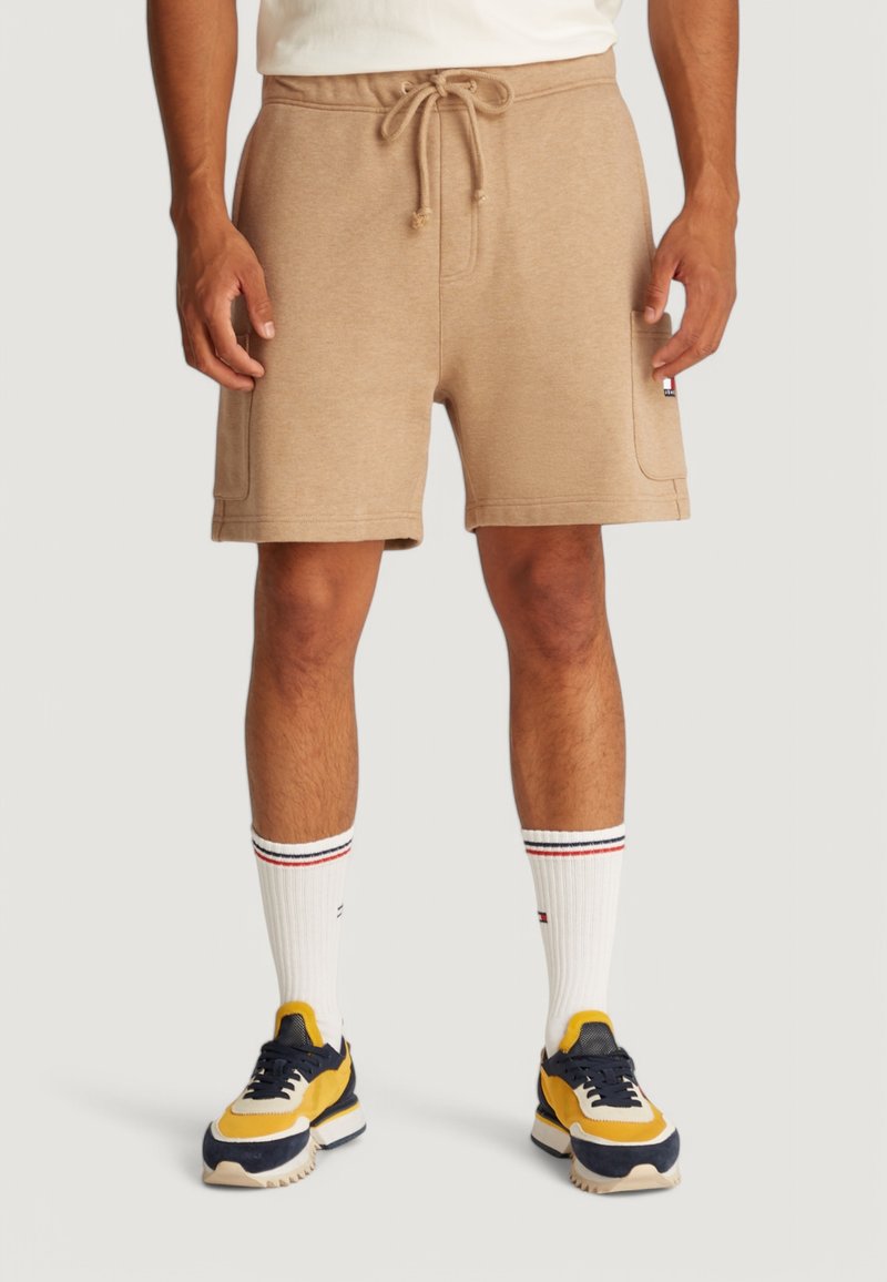 Pantaloni corti con coulisse beige, in tessuto misto cotone, con vita a costine. Abbinati a calzini bianchi con strisce rosse e blu e scarpe da ginnastica colorate.