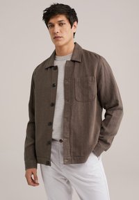 Brown textured button-up jacket with a shirt collar, single chest pocket, and buttons, paired with a light gray sweater and white pants.