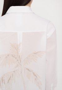 Witte shirt met een doorschijnend achterkantpaneel met een beige palmbomenprint. Klassieke kraag en knoopsluiting.