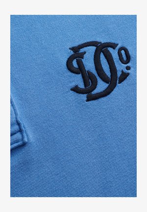 Blauer strukturierter Stoff mit schwarzen gestickten, ineinander verschlungenen Buchstaben und einem kleinen, runden Punktmuster oben links.