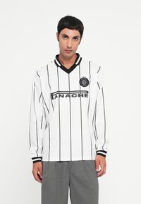 Un jeune homme portant un maillot de sport à manches longues blanc avec des rayures verticales noires et un pantalon gris, se tenant devant un fond blanc uni.