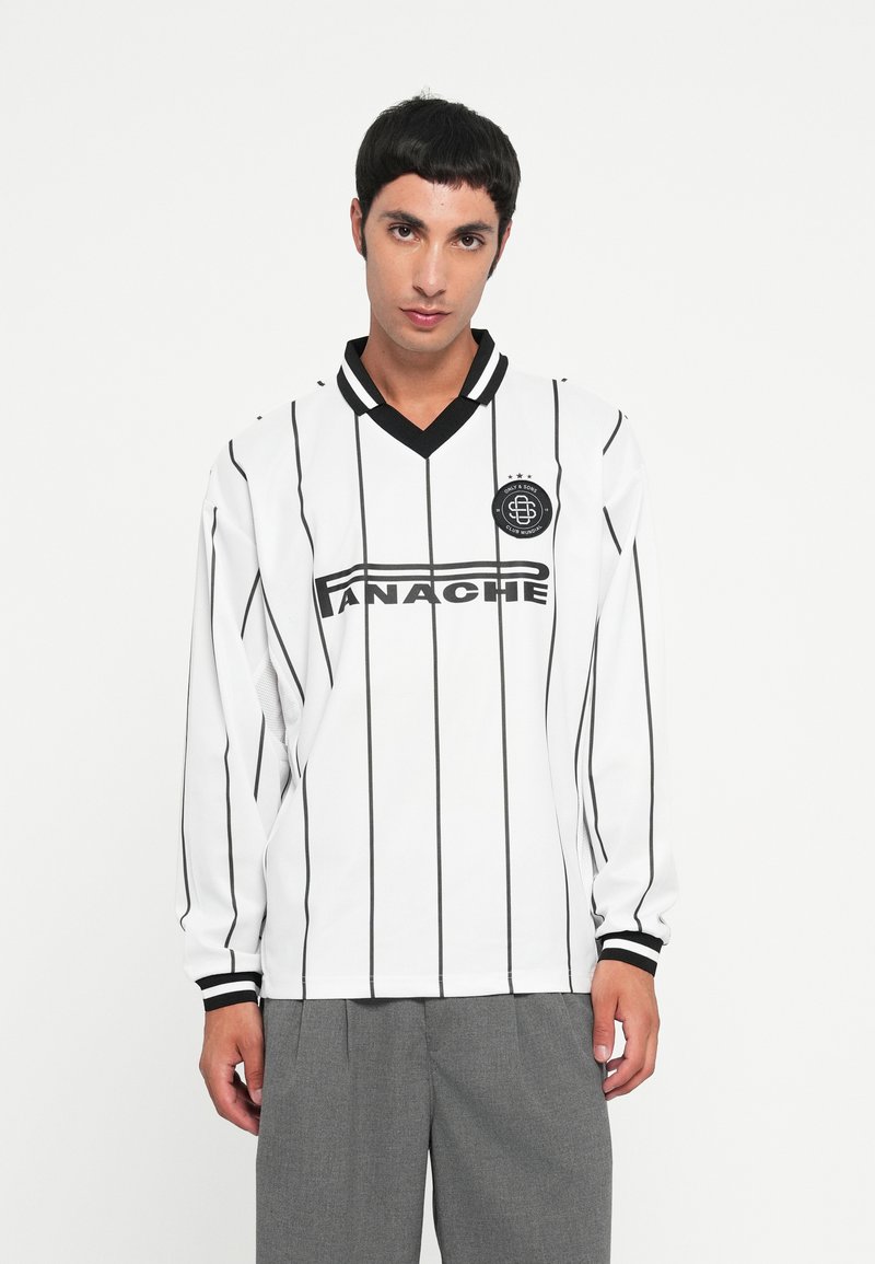 Junger Mann, der ein weißes Sporttrikot mit schwarzen vertikalen Streifen und grauen Hosen trägt, steht vor einem einfachen weißen Hintergrund.