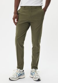 Pantalones de corte ajustado en color verde oliva con un diseño de frente plano, bolsillos laterales y una sutil textura, combinados con zapatillas con acentos metálicos.