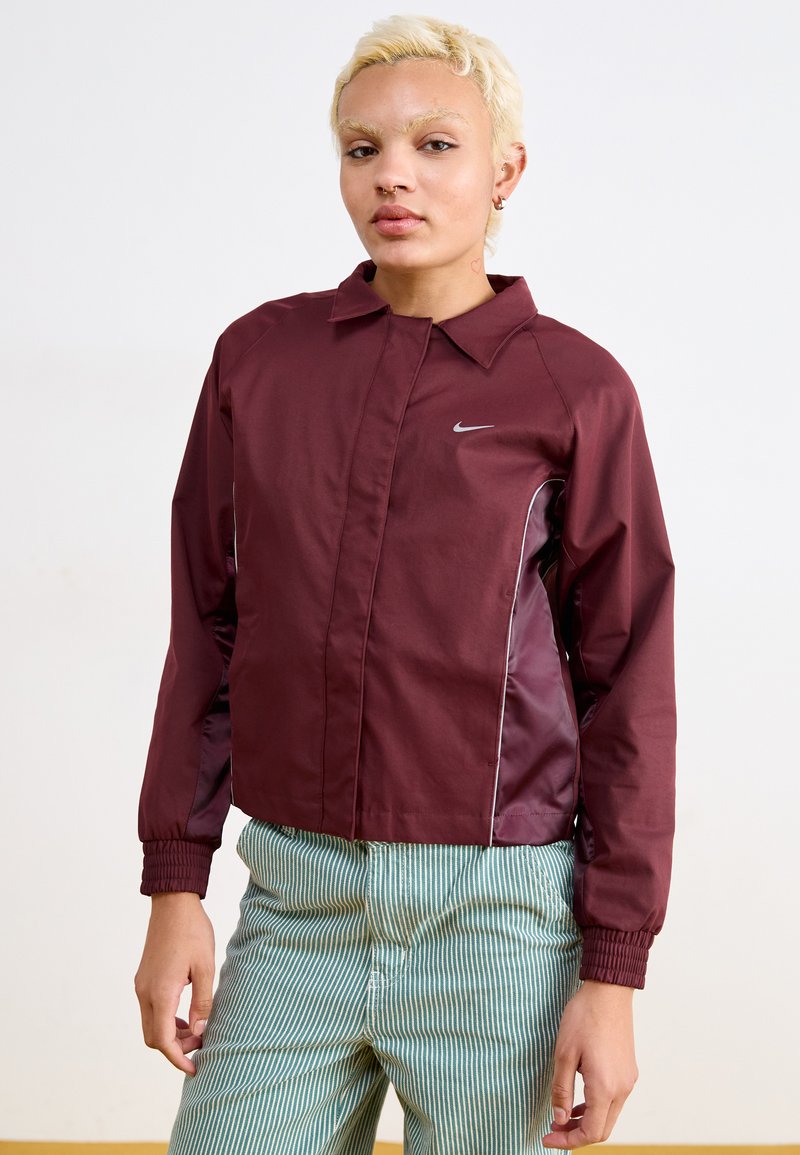 Casaco marrom com colarinho, apresentando um logo prata da Nike e detalhes laterais. Usado com calças verdes listradas. Textura suave.