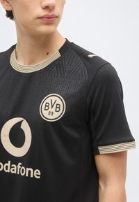 Mlad človek, ki nosi črno nogometno majico Borussia Dortmund z bež logotipi ter črtastim ovratnikom in robovi rokavov.