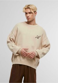 Pulover oversized bej cu mâneci lungi, manșete cu pliuri și un mic logo brodat maro pe piept, asortat cu pantaloni maro.
