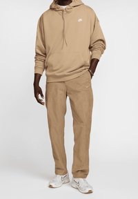 Beige hoodie med en framficka och vit Nike-logotyp, matchande beige byxor och vita sneakers med svarta accenter. Slät textur.