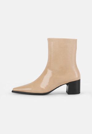 Beige lackläderankelstövlar med spetsig tå och kraftig svart klack. Slät, mjuk textur med en minimalistisk design.