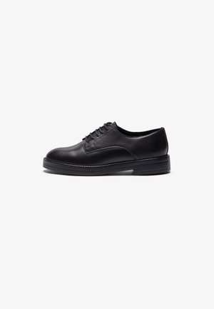 Black leather shoe with a rounded toe, featuring a smooth finish, six eyelets for laces, and a thick black rubber sole.