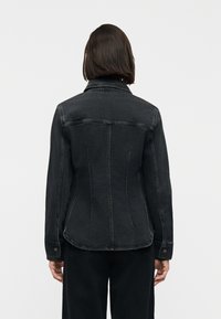 Άτομο που φοράει εφαρμοστό μαύρο τζιν μπουφάν με μακριά μανίκια, φαίνεται από πίσω πάνω σε απλό λευκό φόντο.