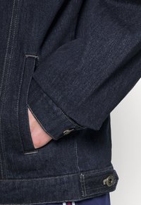 Veste en denim bleu foncé avec une texture lisse, dotée d'un poignet boutonné et d'une poche latérale, mise en valeur par des coutures contrastantes.