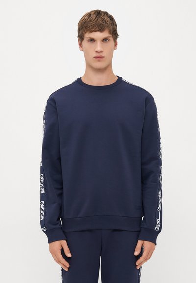Joven vestido con un sudadera de color azul marino y pantalones a juego con rayas blancas del logo de Moschino en las mangas y los lados de los pantalones.