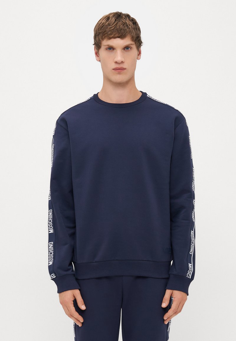 Ung mann i marineblå hettegenser og matchende bukser med hvite Moschino-logo striper på ermene og sidene av buksebenene.