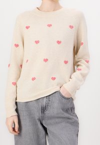 Sweter dzianinowy w kremowym kolorze z różowymi wzorami serc. Okrągły dekolt, ściągacze na rękawach i u dołu, lekko luźny krój. Połączony z jasnoszarymi spodniami.