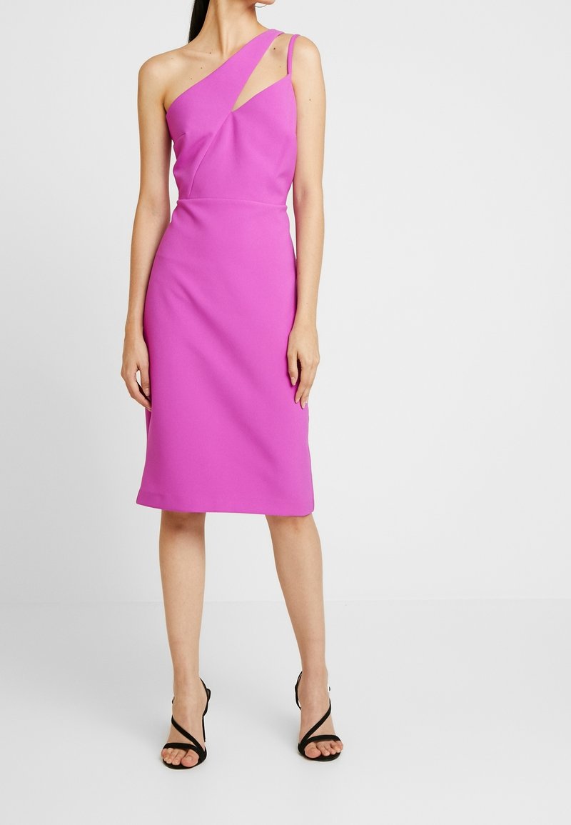 Femme portant une robe rose vif mi-longue à une épaule et des escarpins noirs à bout ouvert, debout sur un sol blanc uni.