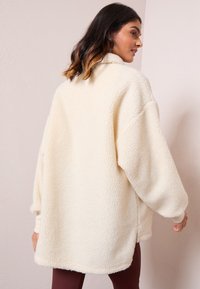 Jachetă din fleece de culoare crem, cu guler înalt, croială oversized și textură moale. Prezintă umeri lăsați și manșete ribbate.