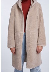 Osoba v dlouhém béžovém teddy kabátu s kapucí, oblečená ve spadeném hnědém roláku a světle modrých roztržených džínách, stojící na jednolitém pozadí.