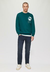 Teal Sweatshirt mit Mondgrafik und weißem Text auf der Brust, kombiniert mit dunklen Jeans und beigen Sneakers. Glatter Stoff, lässige Passform.
