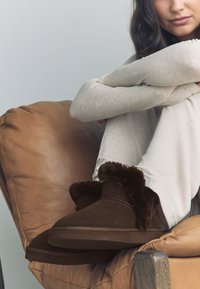 Hnědé semišové pantofle s huňatým kožíškovým lemováním. Měkká textura a zaoblený design špičky. Nosit na nohou osoby proti hnědému koženému křeslu.