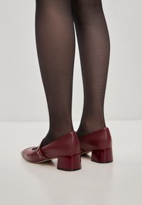 Burgundin nahasta valmistetut lohkarekorkoiset kengät, joissa on pyöreä kärki ja sivupanta, yhdistettynä mustiin läpinäkyviin sukkahousuihin. Silkkinen pinta, matalaleikkauksinen muoto.