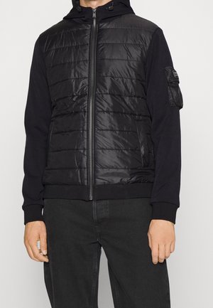 Mann trägt eine schwarze gesteppte Jacke mit Reißverschluss und Kapuze, mit gerippten Bündchen und einer Tasche am linken Ärmel, kombiniert mit schwarzen Hosen.