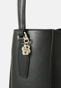 Sac à main en cuir noir avec une finition lisse, équipé d'un charm doré et d'une anse cousue. Base carrée et forme structurée.