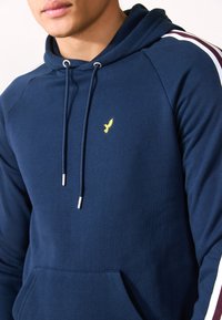 Sudadera azul marino hecha de suave algodón, con un bolsillo frontal, cordones ajustables y un logo dorado de águila en el pecho.