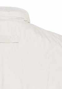 Quiltet lys beige jakke med høj krave, der har diskrete buede syninger og et logo-tag nær halsudskæringen.