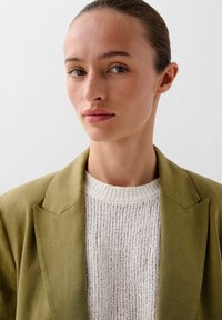 Olive green blazer with notched lapels over a cream, textured knit sweater. The garment features a tailored fit, emphasizing a structured silhouette.