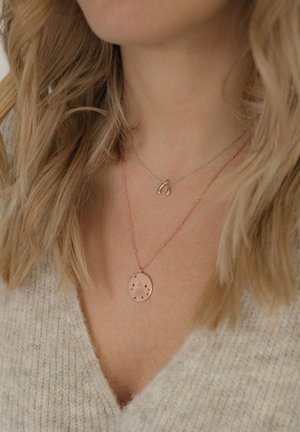Schichtkette: Eine silberne Kette mit einem Wunschnachtschädel-Anhänger und einem roségoldenen Anhänger mit einem Baumdesign und Sternen, getragen über einem beigen Pullover.