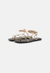 Sandales à talons strappy en fil doré avec une semelle plate noire, présentant un design minimaliste, un bout ouvert et une sangle de cheville ajustable.