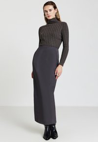 Maglione dolce grigio scuro a costine con collo alto e gonna lunga e strutturata dalla silhouette aderente. Modella che indossa stivali neri alla caviglia. Sfondo neutro.