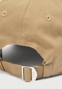 Khakin värinen lippalakki, jossa on teksturoitu kangas, kaareva lippa ja takana metallinen solkikiinnitys koon säätämiseksi.