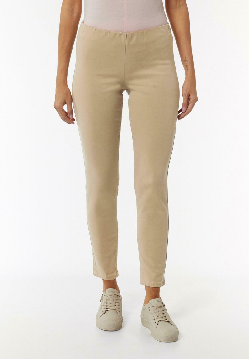 Beige stretchige Hosen mit einem figurschmeichelnden Design, schmal zulaufendem Bein und glatter Textur. Kombiniert mit hellen Sneakers. Minimale Details.
