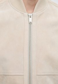 Κοντινή λήψη μιας μπεζ σουέντ ζακέτας με ασημένιο φερμουάρ και ribbed γιακά, μερικώς φερμουαρισμένη στο κέντρο μπροστά.