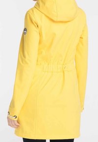 Gul huva jacka med insnörd midja, slät tyg och logotyp på vänster ärm. Har långa ärmar och en figursydd silhuett.