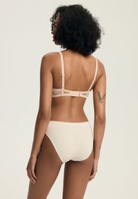 Kobieta z kręconymi włosami, w beżowym koronkowym biustonoszu i pasującym komplecie bielizny, stojąca bokiem z tyłem do aparatu, pokazująca tatuaż motyla na ramieniu.